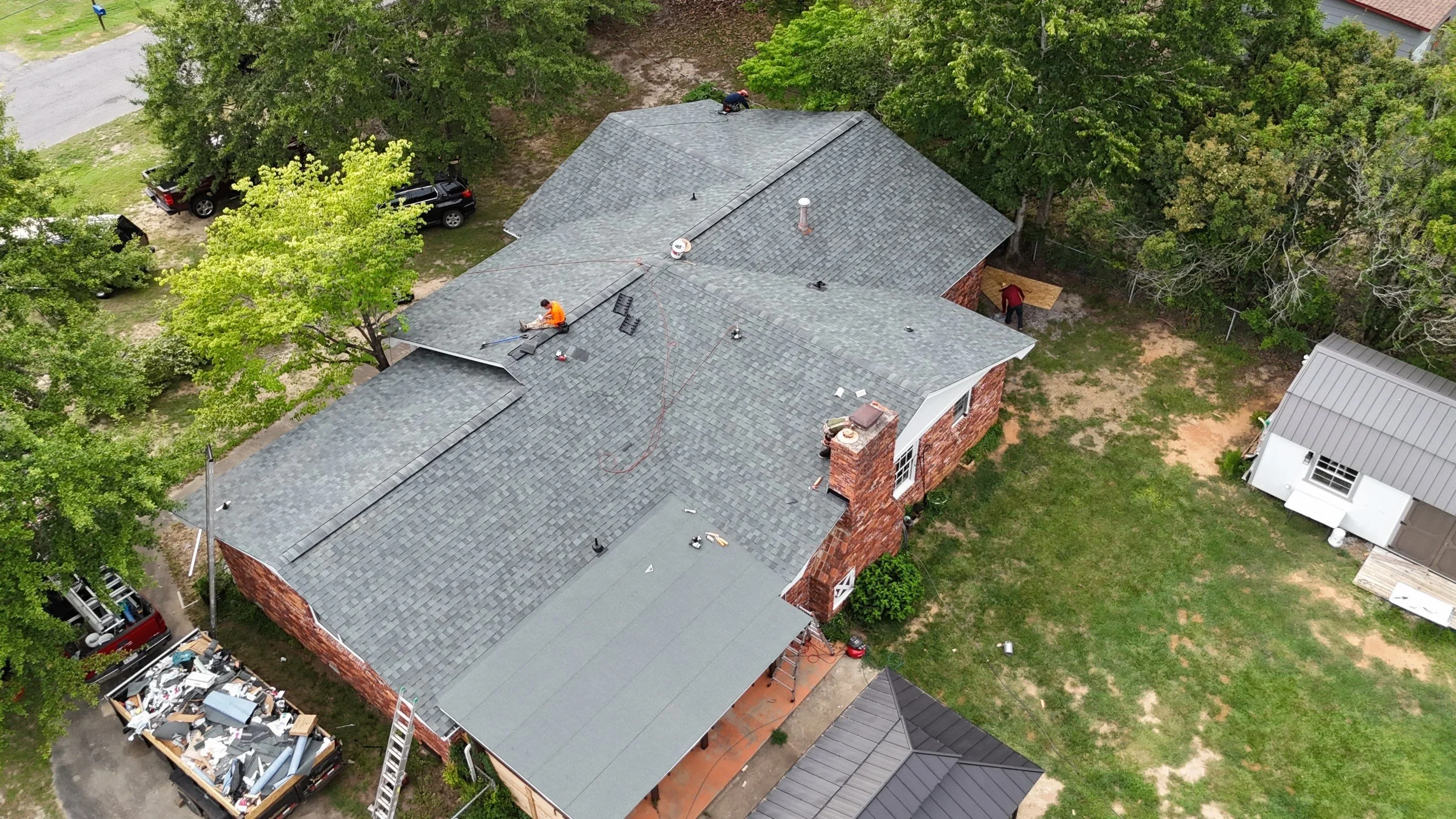 Gray asphalt shingle roof installation on brick home with workers and safety equipment