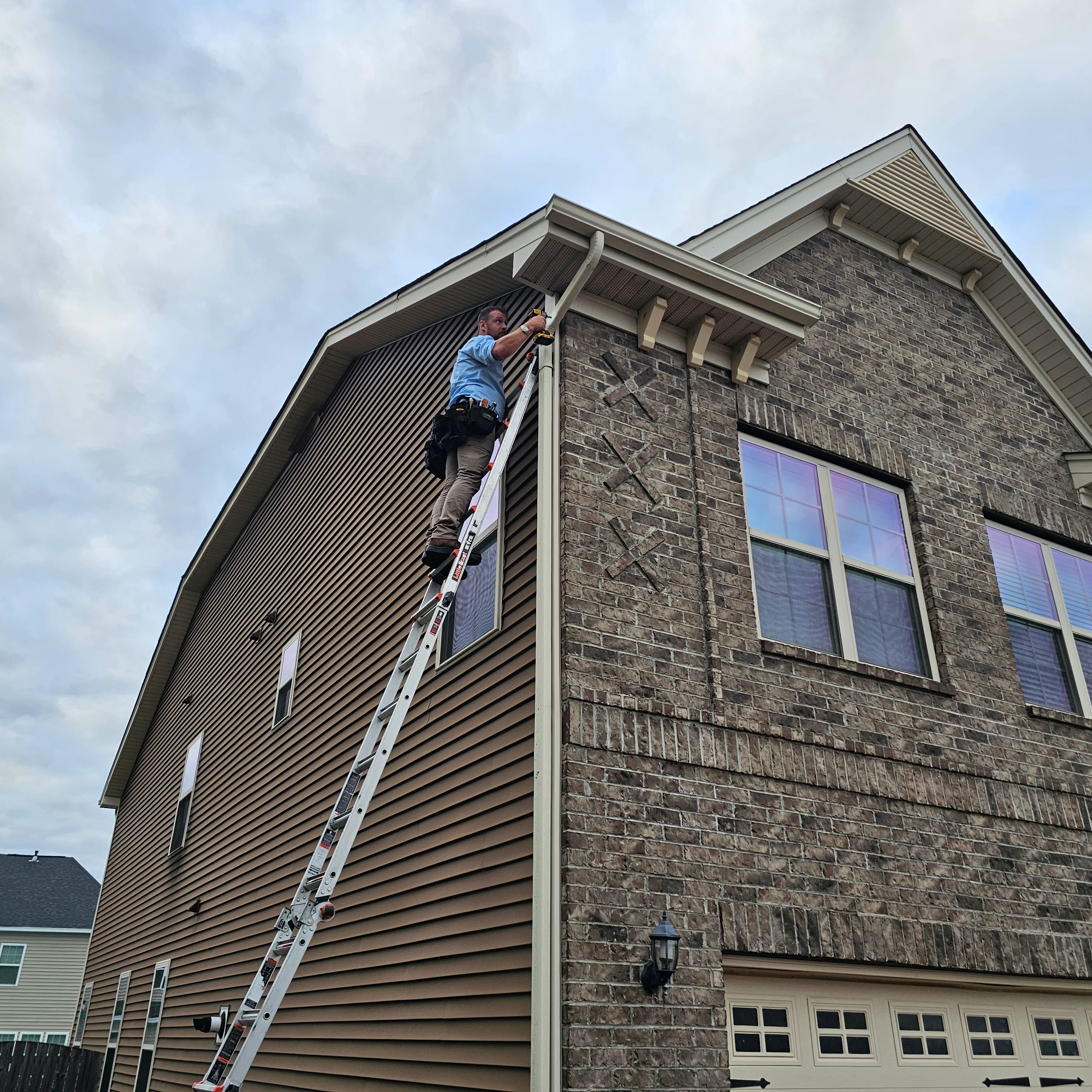 Worker on ladder installing gutters on two-story home with vinyl siding and stone veneer
