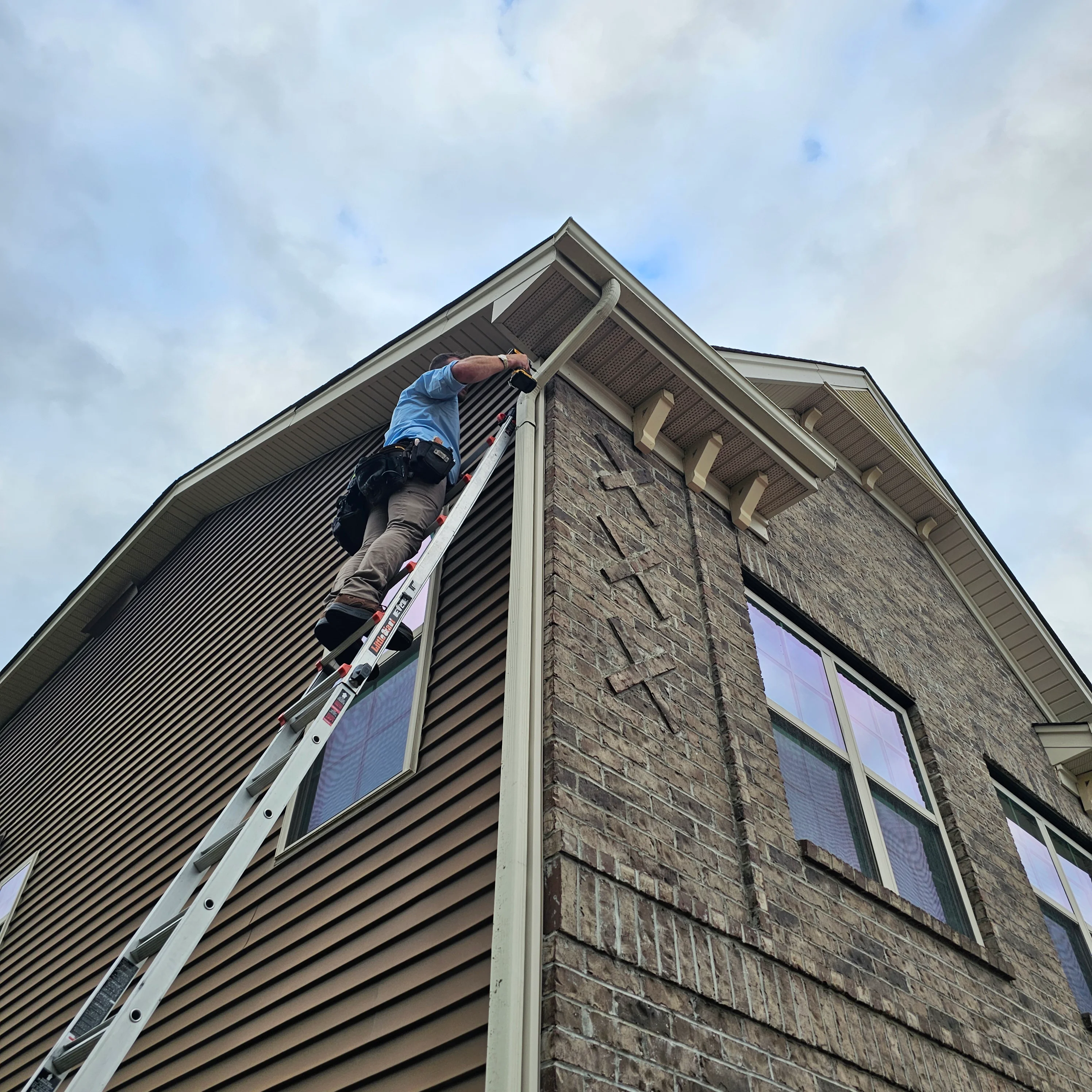 Worker on ladder installing gutters and soffit on two-story home with stone and vinyl siding